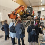 Kuvassa Palosuojelurahaston sihteeristö seisoo Pirkanmaan VPK:n kalustohallissa telakaivukoneen edessä ja kuuntelee Henrik Joen esitelmää VPK:n kalustosta.