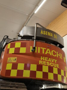Vesisäiliössä lukee asema 55. Telakaivukoneen takaosa on kuvioitu puna-keltaisin huomiovärein. Takaosassa lukee Hitachi heavy rescue.