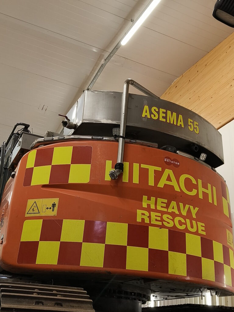 Vesisäiliössä lukee asema 55. Telakaivukoneen takaosa on kuvioitu puna-keltaisin huomiovärein. Takaosassa lukee Hitachi heavy rescue. 