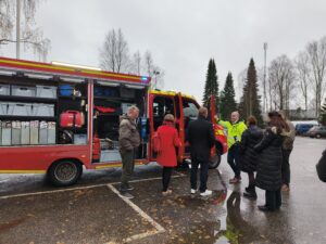 Pelastusauto on paloaseman parkkipaikalla. Ulkona sataa vettä. Pelastusauton ovet ovat auki. Palosuojelurahaston hallitus ja sihteeristö tarkastelee auton varustelua ja kuuntelee Lindbergin esittelyä.