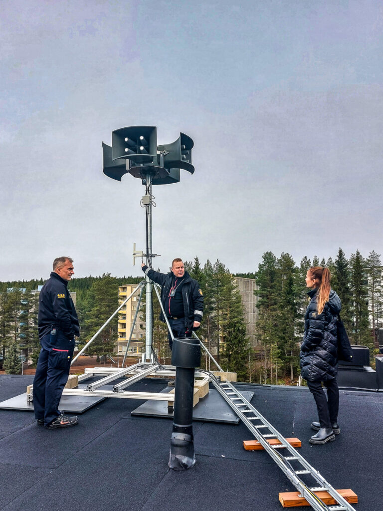 Herrala, Karilahti ja Hyvärinen ovat talon katolla suurtehohälyttimen luona. Suurtehohälyttimestä lähtee kaiuttimet neljään ilmansuuntaan. Taustalla näkyy kerrostaloja ja metsikköä.