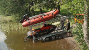 Kaksikerroksinen kuljetustraileri rantaveteen kallistettuna. Trailerin yläkerroksessa on moottorikumivene ja alakerroksessa vesijetti. Henkilö liu’uttaa vesijettiä rantaveteen trailerilta.