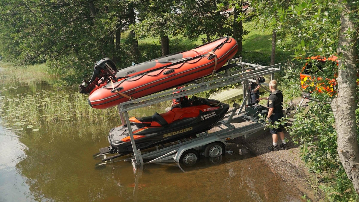 Kaksikerroksinen kuljetustraileri rantaveteen kallistettuna. Trailerin yläkerroksessa on moottorikumivene ja alakerroksessa vesijetti. Henkilö liu’uttaa vesijettiä rantaveteen trailerilta.